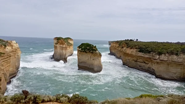 A natural rock archway, known as the Island Archway, stands majestically in the ocean, with waves crashing against its base and a sandy beach visible. It's part of the iconic Great Ocean Road coastline.
