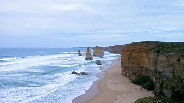 A dramatic coastal landscape featuring towering sea stacks rising from the turbulent turquoise waters of the ocean, with waves crashing onto a sandy beach and steep cliffs lining the shore under an overcast sky.